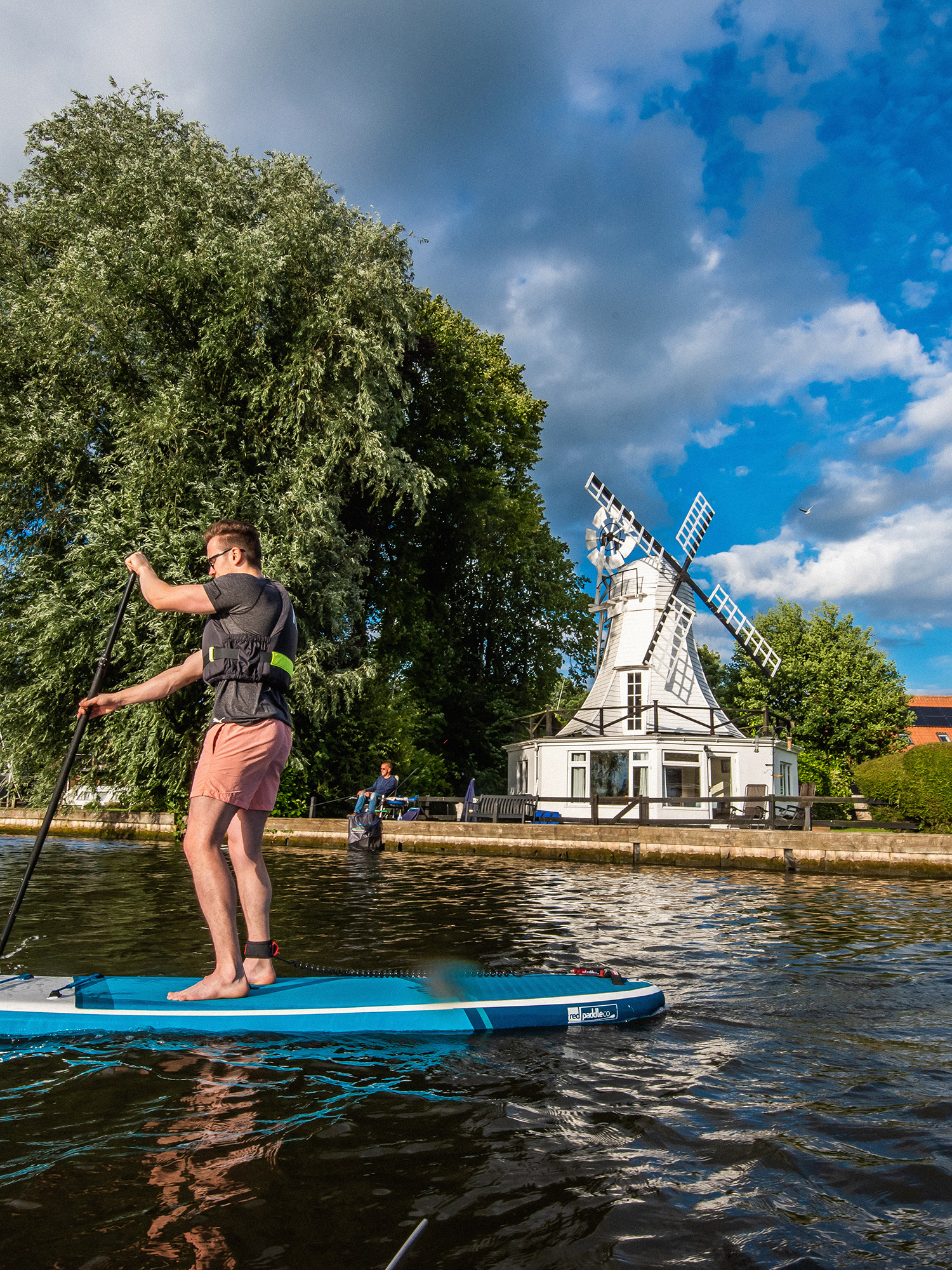 A person paddleboarding past a white wooden chalet with a windmill on top. It's a sunny day and the trees are green with leaves.