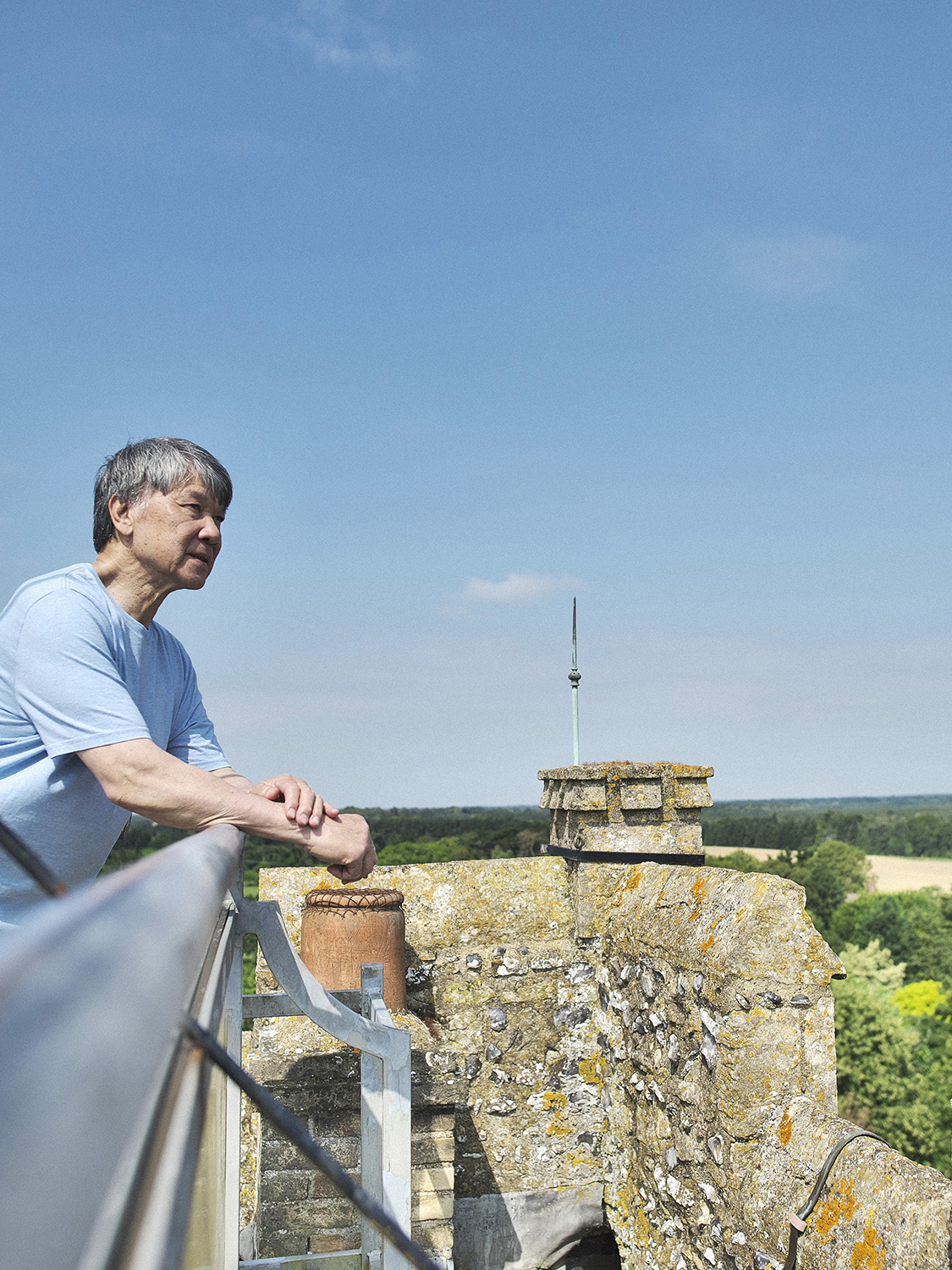 A man wearing a light blue t shirt stands on the edge of a church tower looking across the landscape