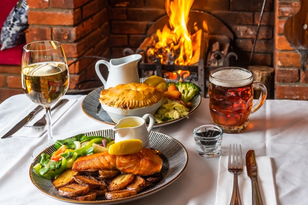 Two large plates of food, a glass of wine and a pint of beer, on a table in front of an open fire